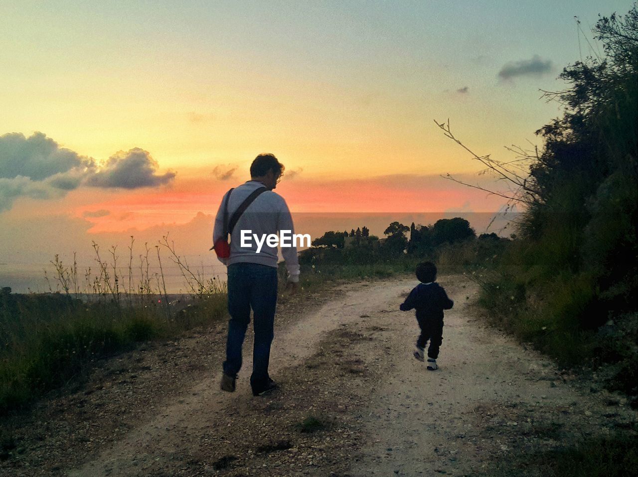 Rear view of father and son walking on dirt road against sky during sunset