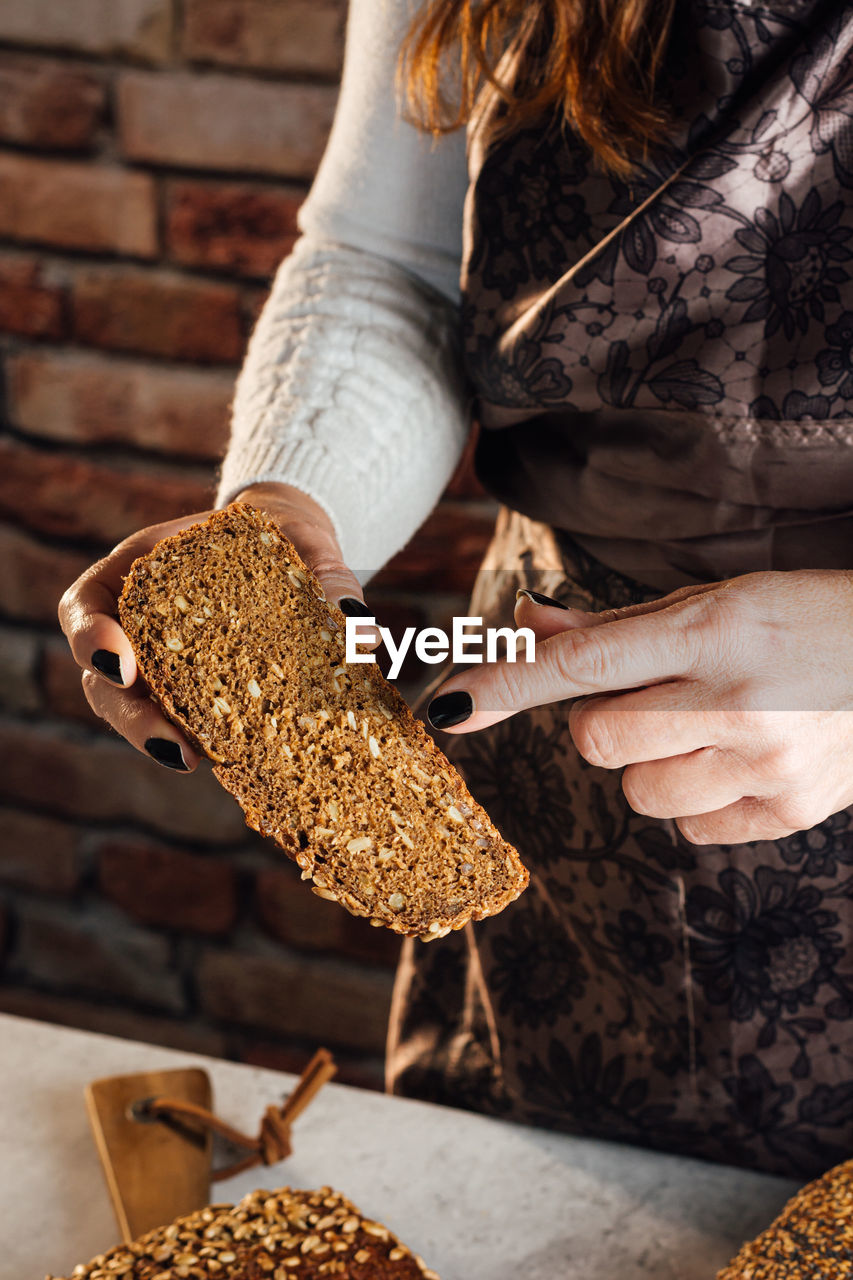 Crop anonymous female baker demonstrating soft fresh bread with crunchy seeds at table in bakery