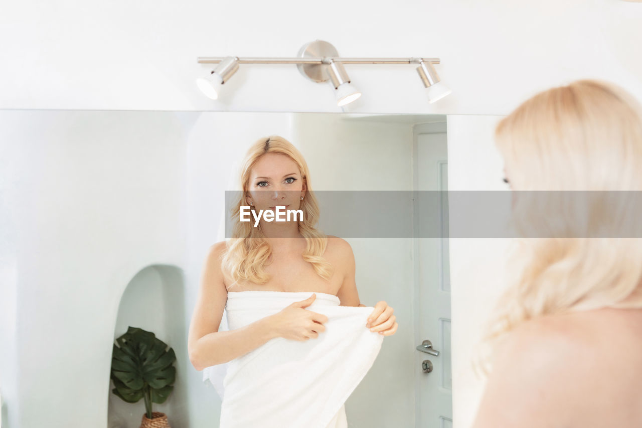 Blond woman in a white bathroom looking at her reflection in the mirror, wearing a towel