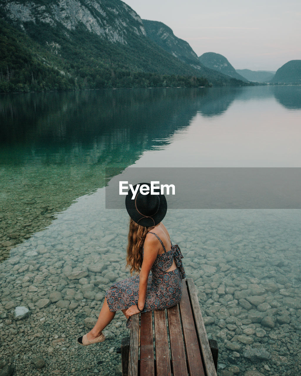 Side view of woman sitting on pier over lake against mountains and sky