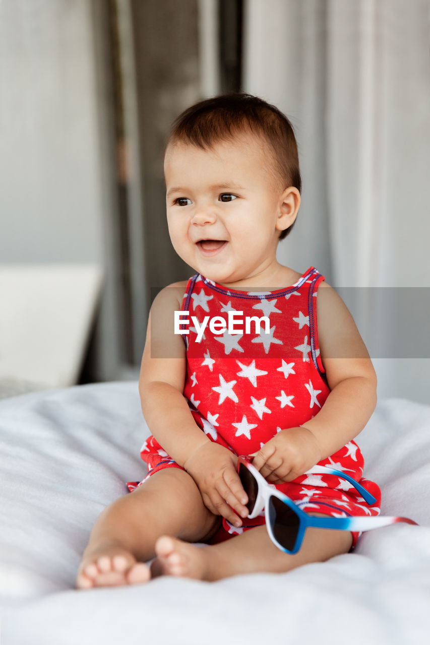 Cute baby girl holding sunglasses while sitting on bed at home