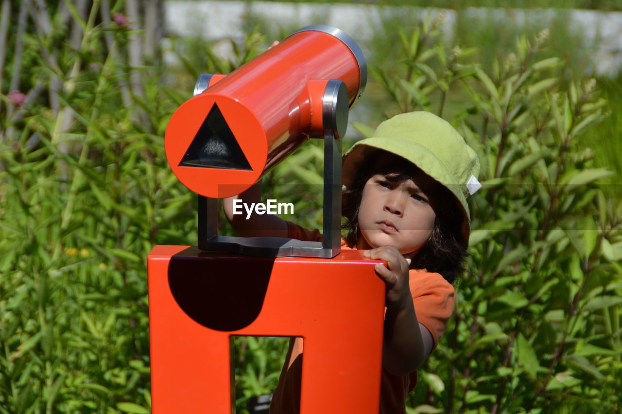 Boy using red hand-held telescope against plants on sunny day