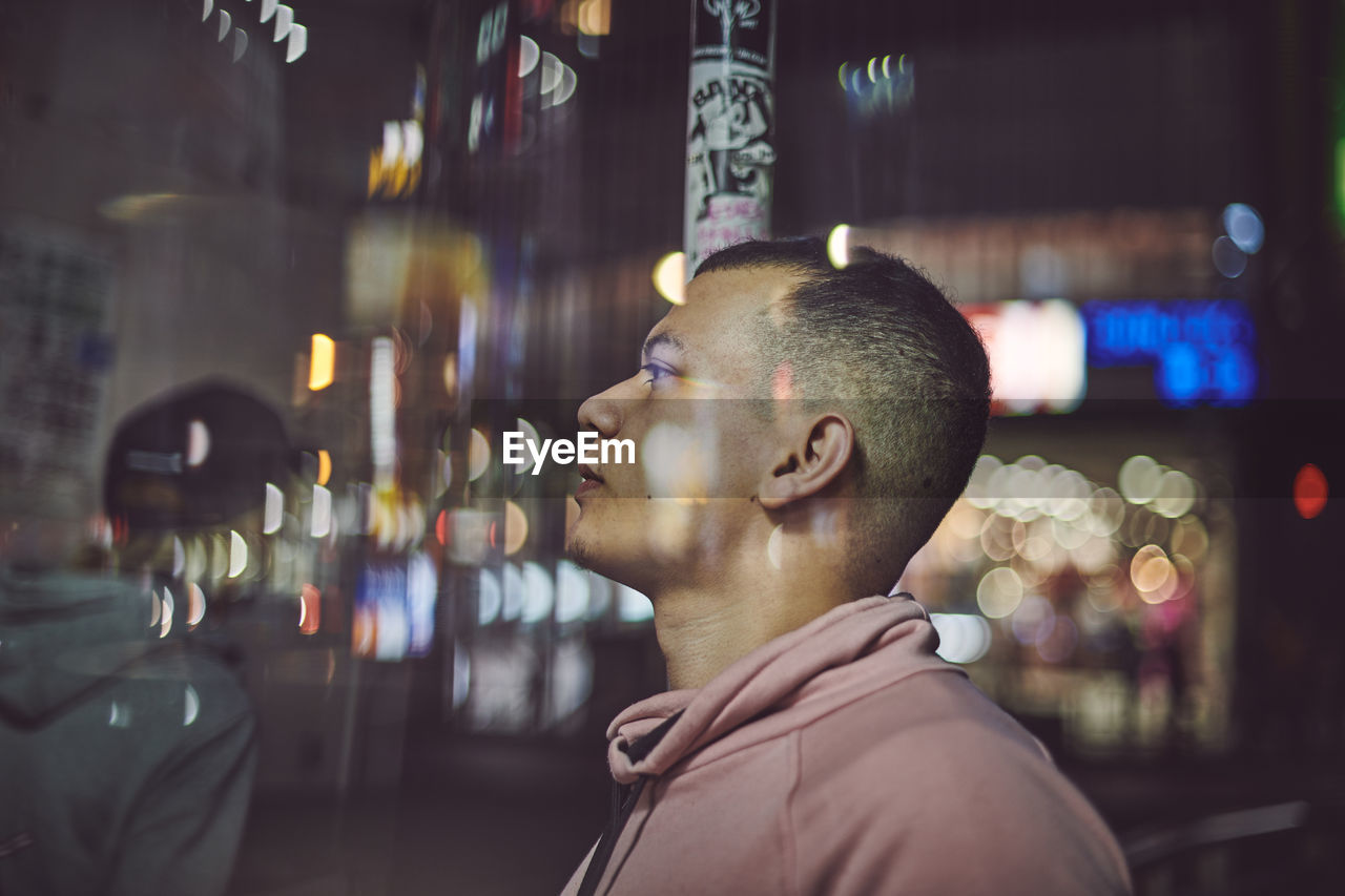 Portrait of young man looking at illuminated city at night with lots of reflections and bokeh