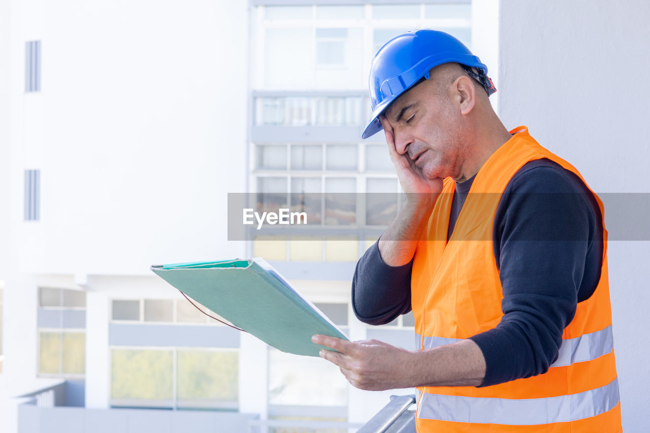 hard hat, adult, occupation, working, industry, helmet, technology, headwear, men, business, one person, hat, construction industry, reflective clothing, protection, engineering, looking, business finance and industry, standing, planning, architecture, holding, computer, manual worker, protective workwear, wireless technology, examining, construction site, person, building contractor, digital tablet, waist up, clothing, communication, construction worker, security, plan, indoors, side view, mature adult, blueprint, expertise, built structure, professional occupation, building, looking down, copy space, occupational safety and health, reading, quality control