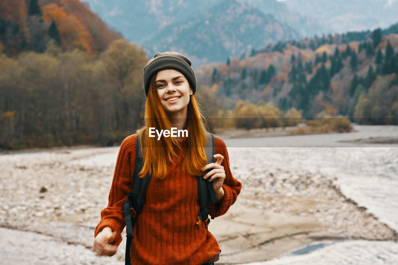 PORTRAIT OF SMILING YOUNG WOMAN STANDING AGAINST MOUNTAIN
