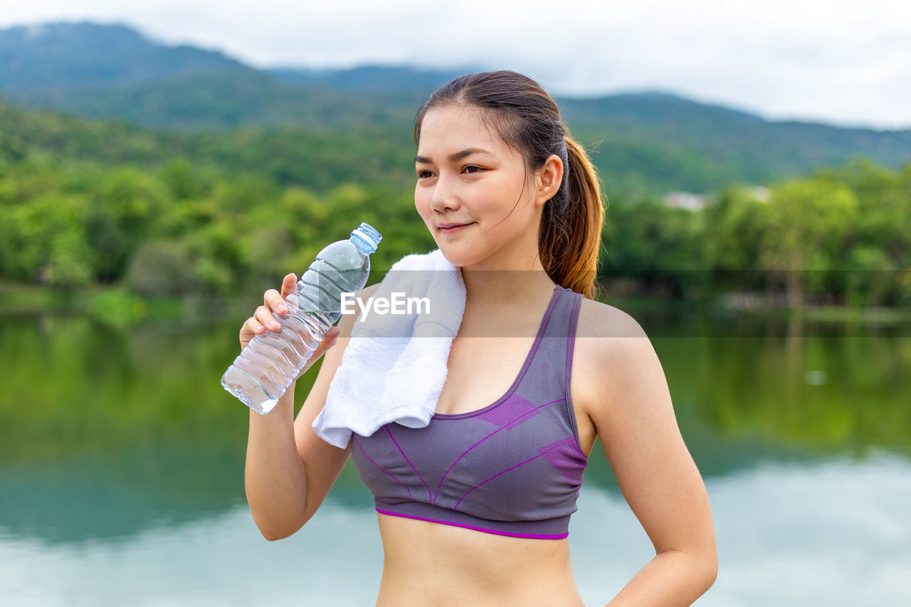 YOUNG WOMAN DRINKING WATER FROM BOTTLE WHILE STANDING AT SHORE