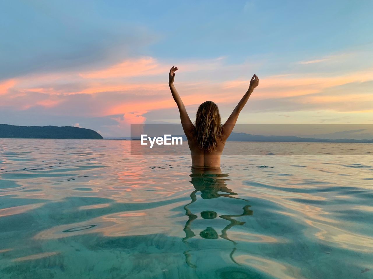 REAR VIEW OF MAN WITH ARMS RAISED IN SEA AGAINST SKY