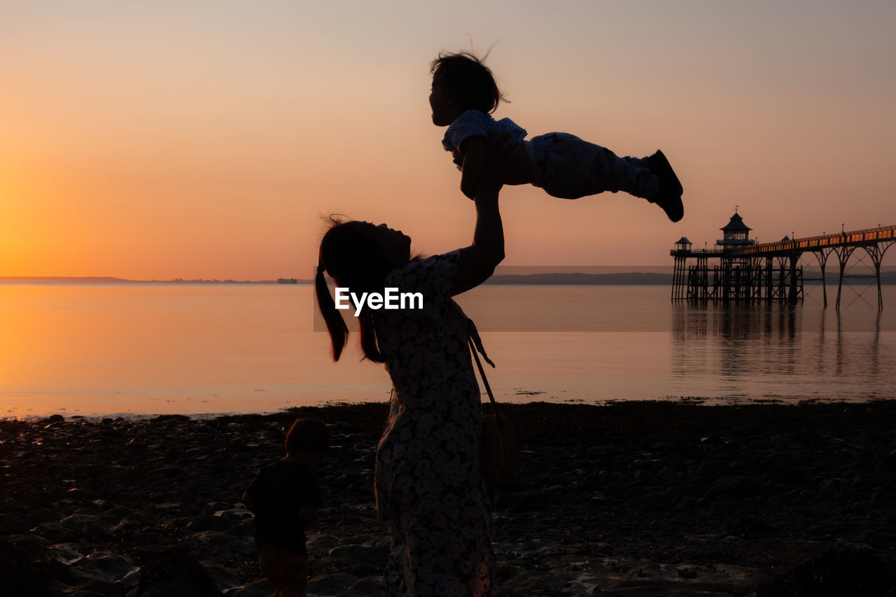 Silhouette woman standing on beach with toddler against sky during sunset