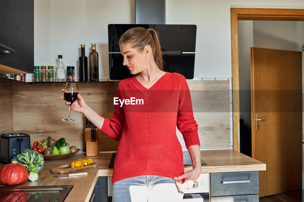 A young girl stands in the kitchen and is distracted from cooking drinking wine from a glass
