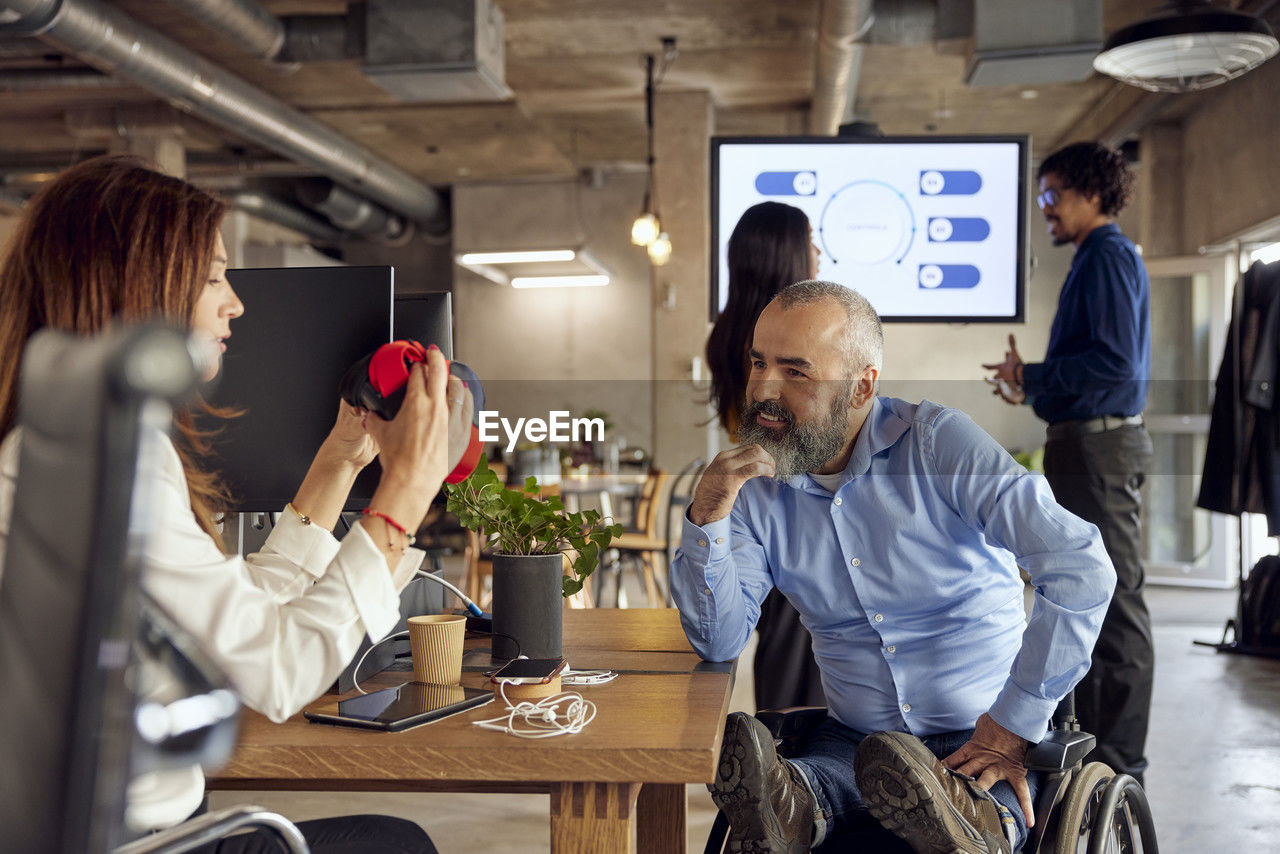 Smiling businessman with disability looking at colleague discussing over vr simulator in creative office