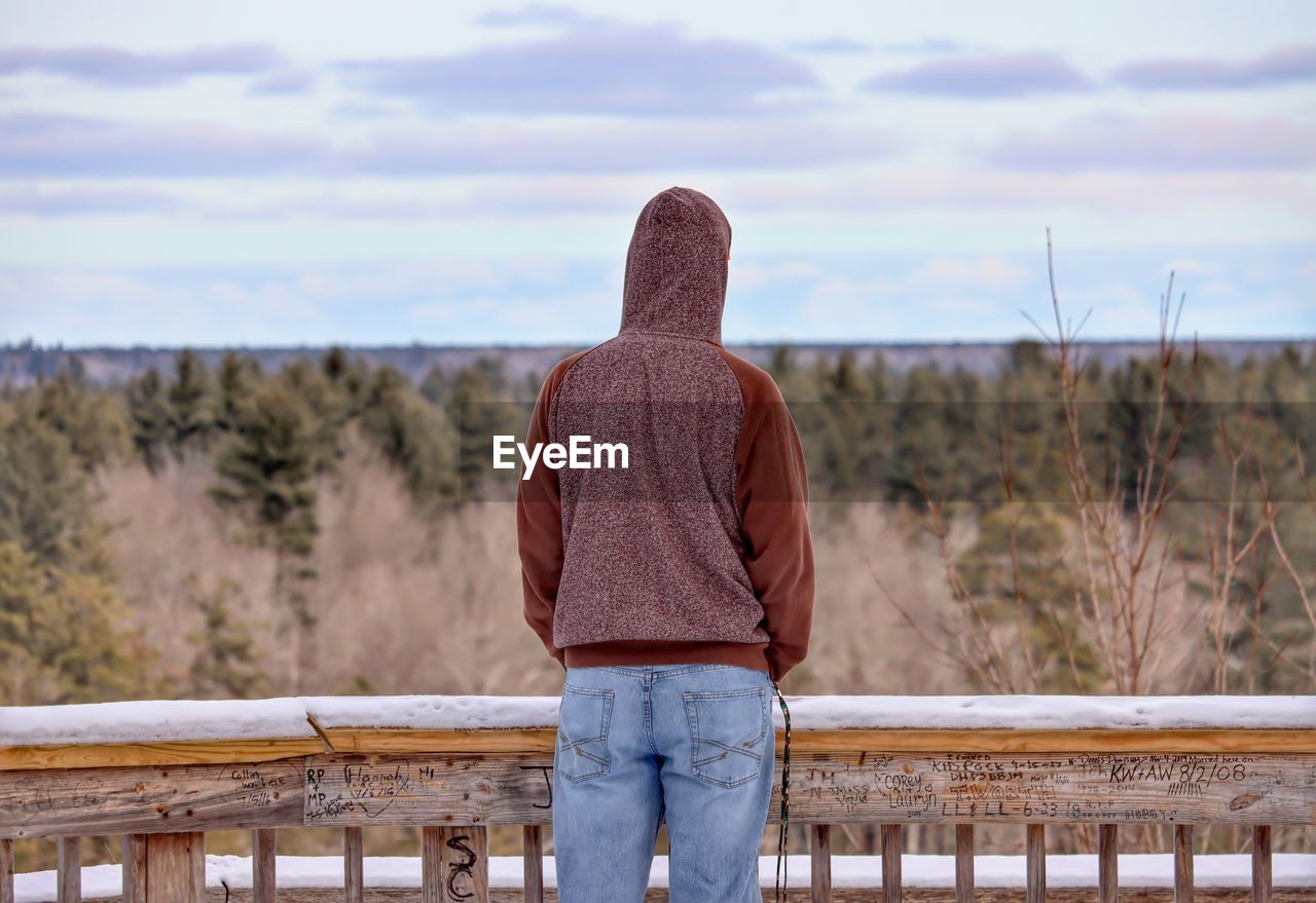 Rear view man wearing hood standing by railing while looking at forest against sky