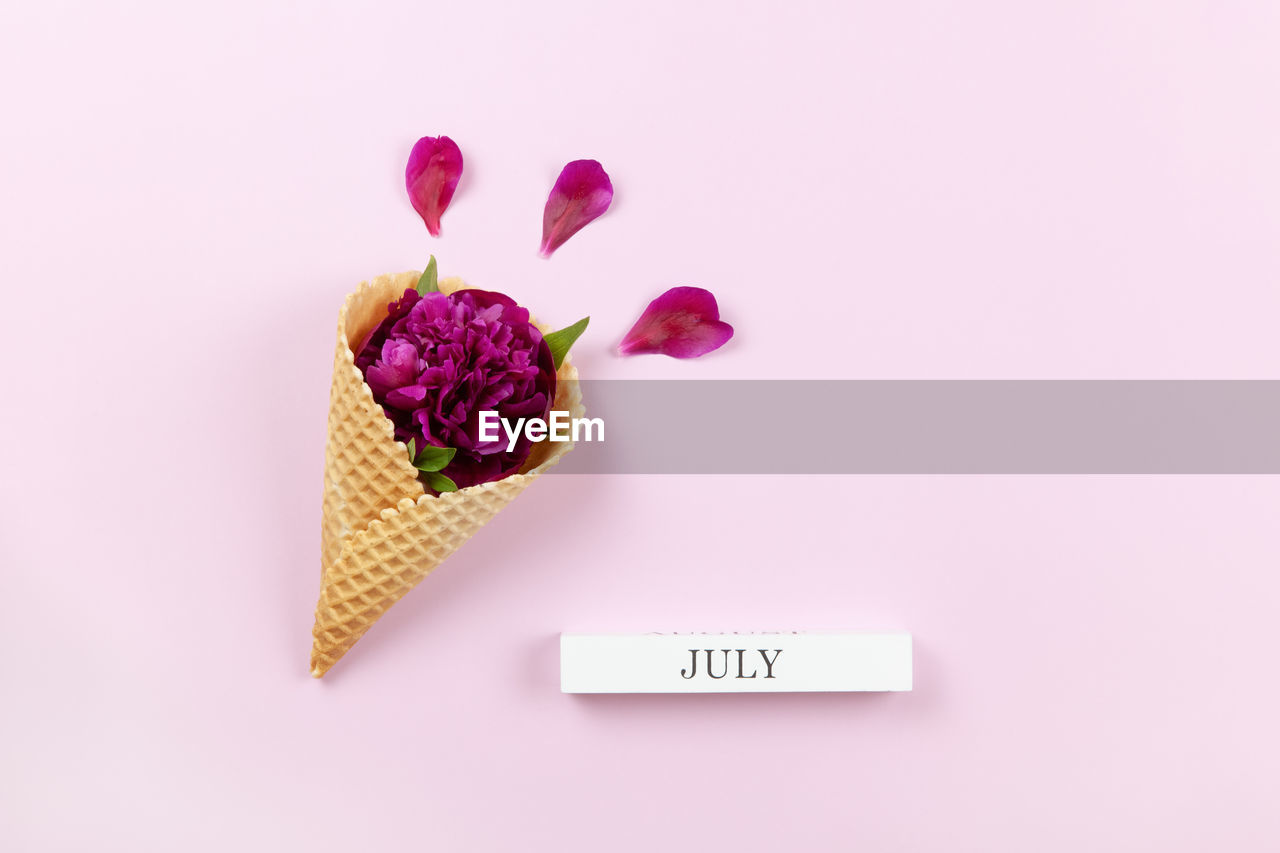 Peony flower in waffle cone, petals, wooden block with inscription july on pastel lilac background
