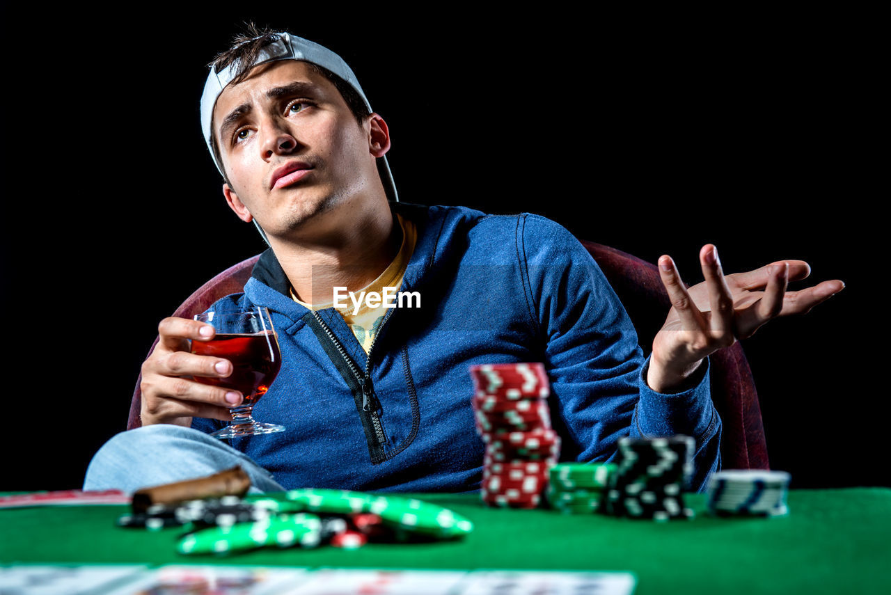Sad man sitting at poker table with whiskey against black background