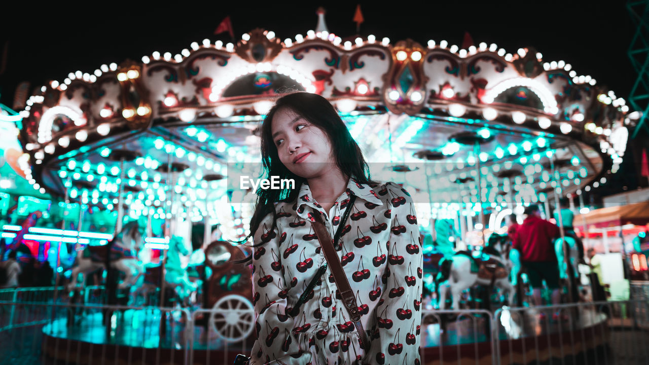 PORTRAIT OF YOUNG WOMAN STANDING AT ILLUMINATED CAROUSEL