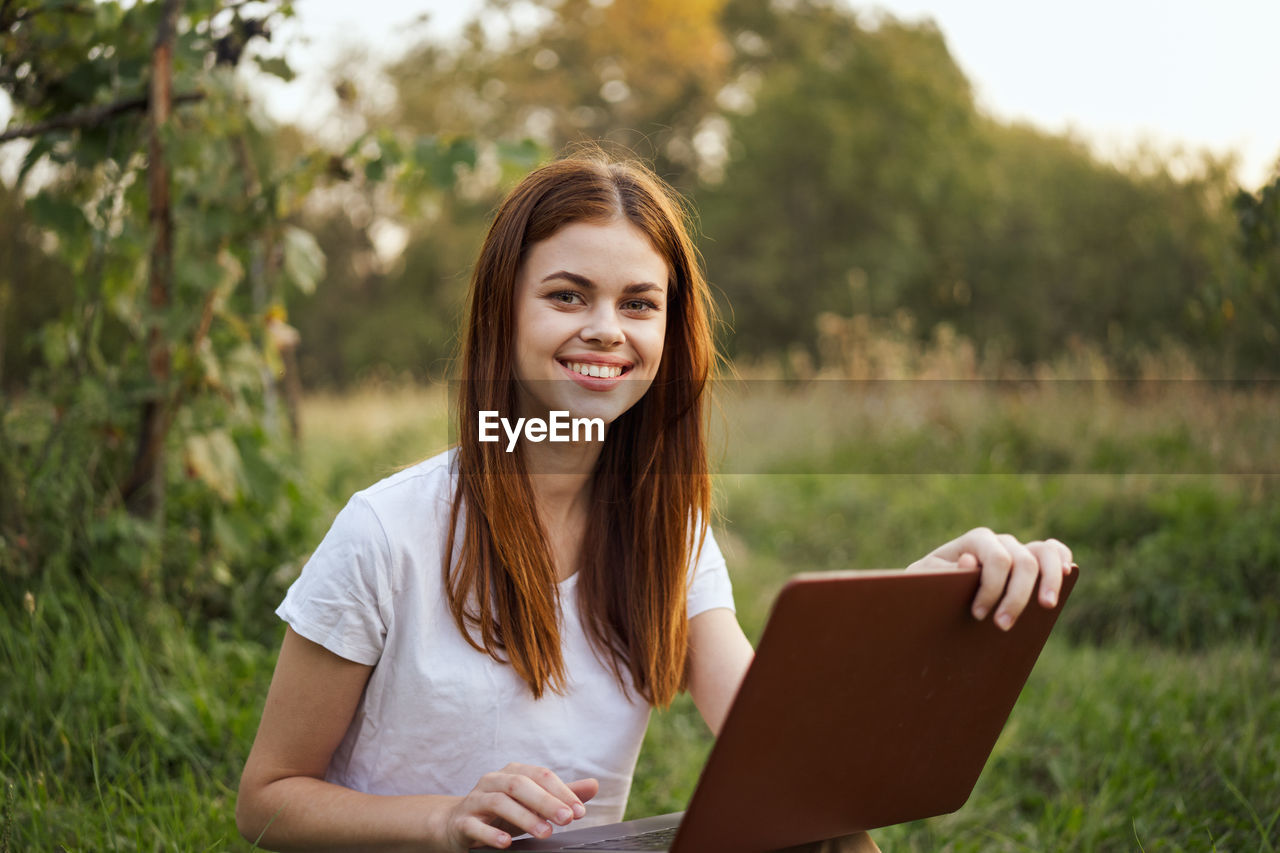 portrait of smiling young woman using laptop while standing against trees