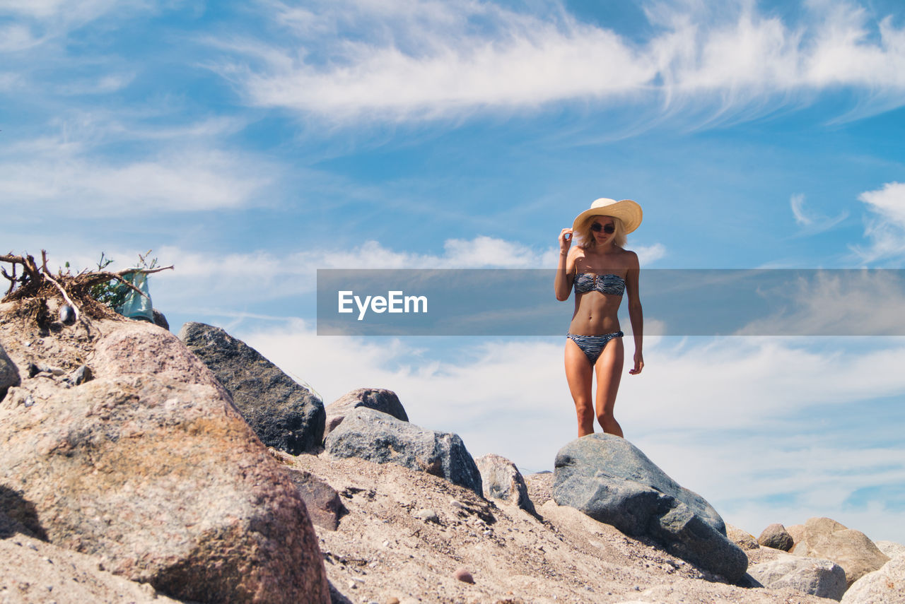 Low angle view of young woman in bikini walking on rock against sky