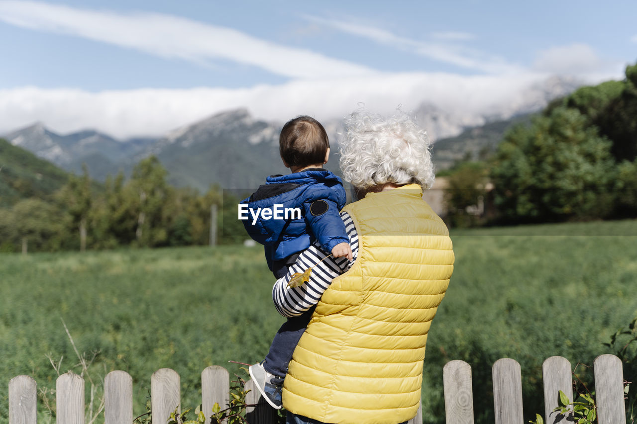 Rear view of a grandmother wearing a medical mask carrying her little grandson in a field