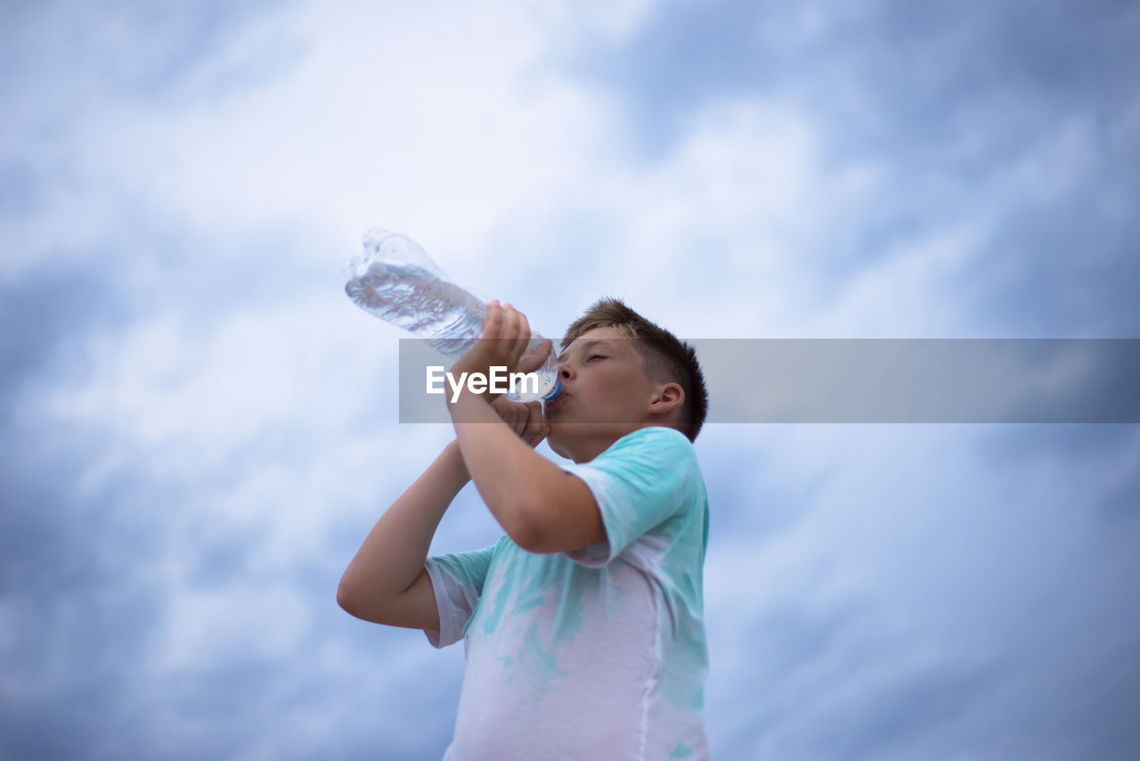 A boy against the sky drinks water from a plastic bottle. thirst. heat. cloudy sky.
