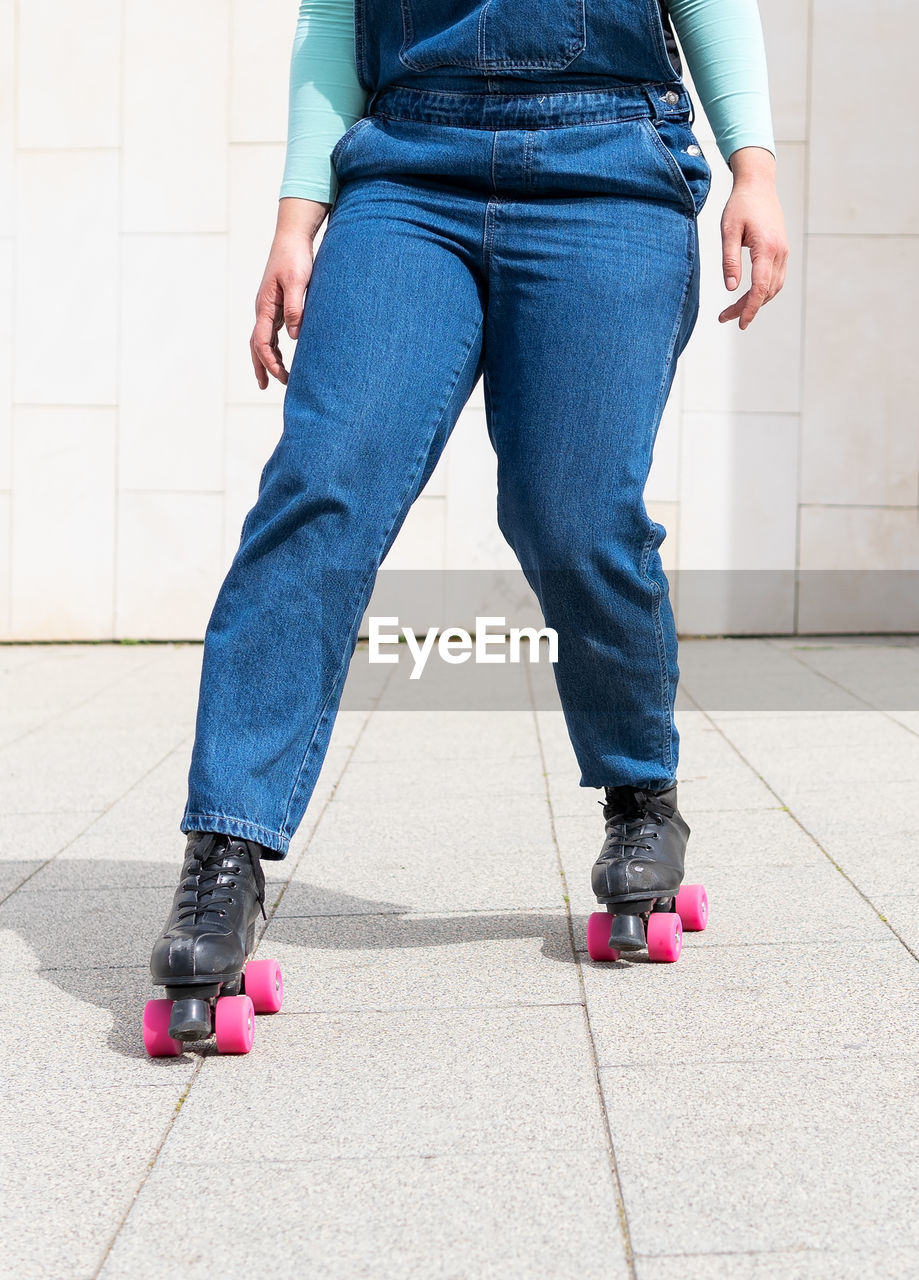 Cropped unrecognizable lady with short hair in denim overall riding roller skates on pavement against building wall