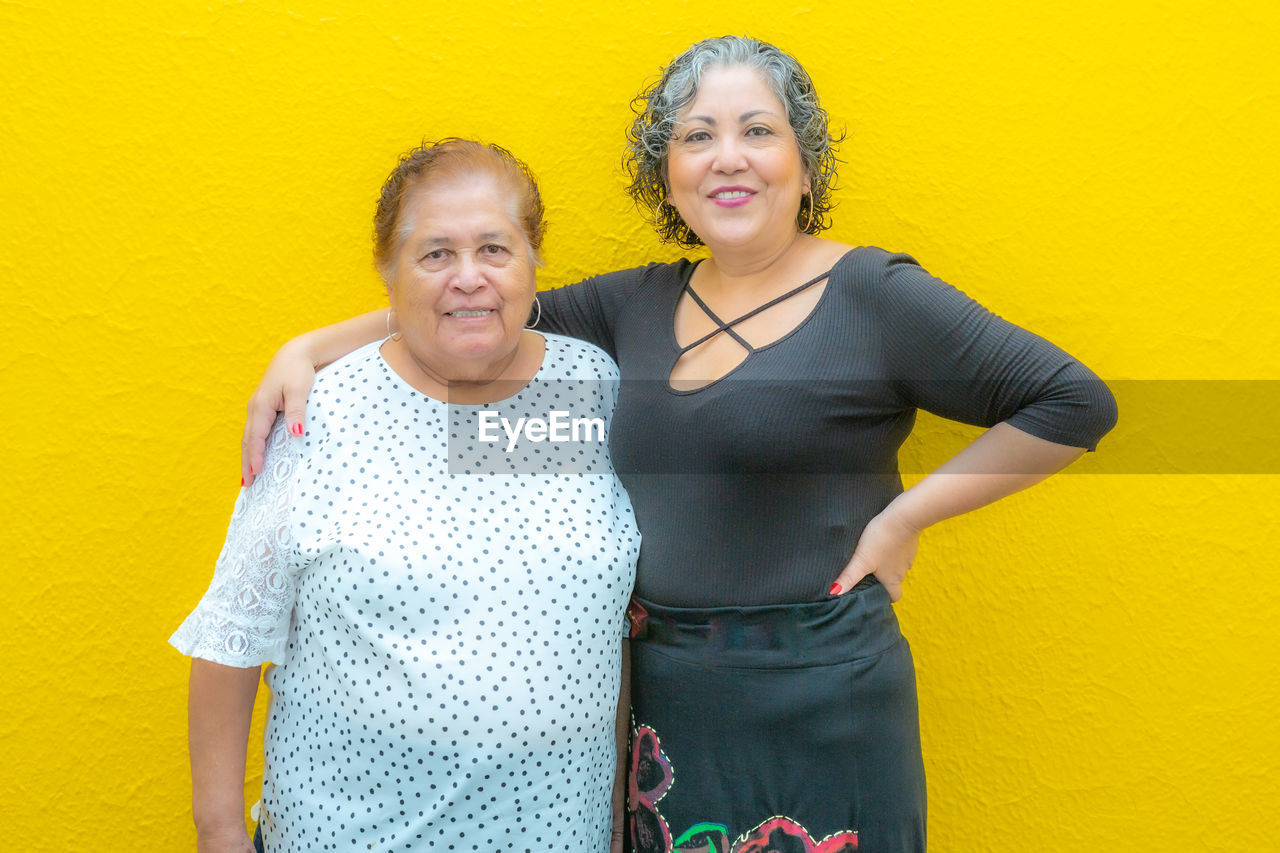 Waist up portrait of a daughter hugging her smiling mother, standing against yellow background