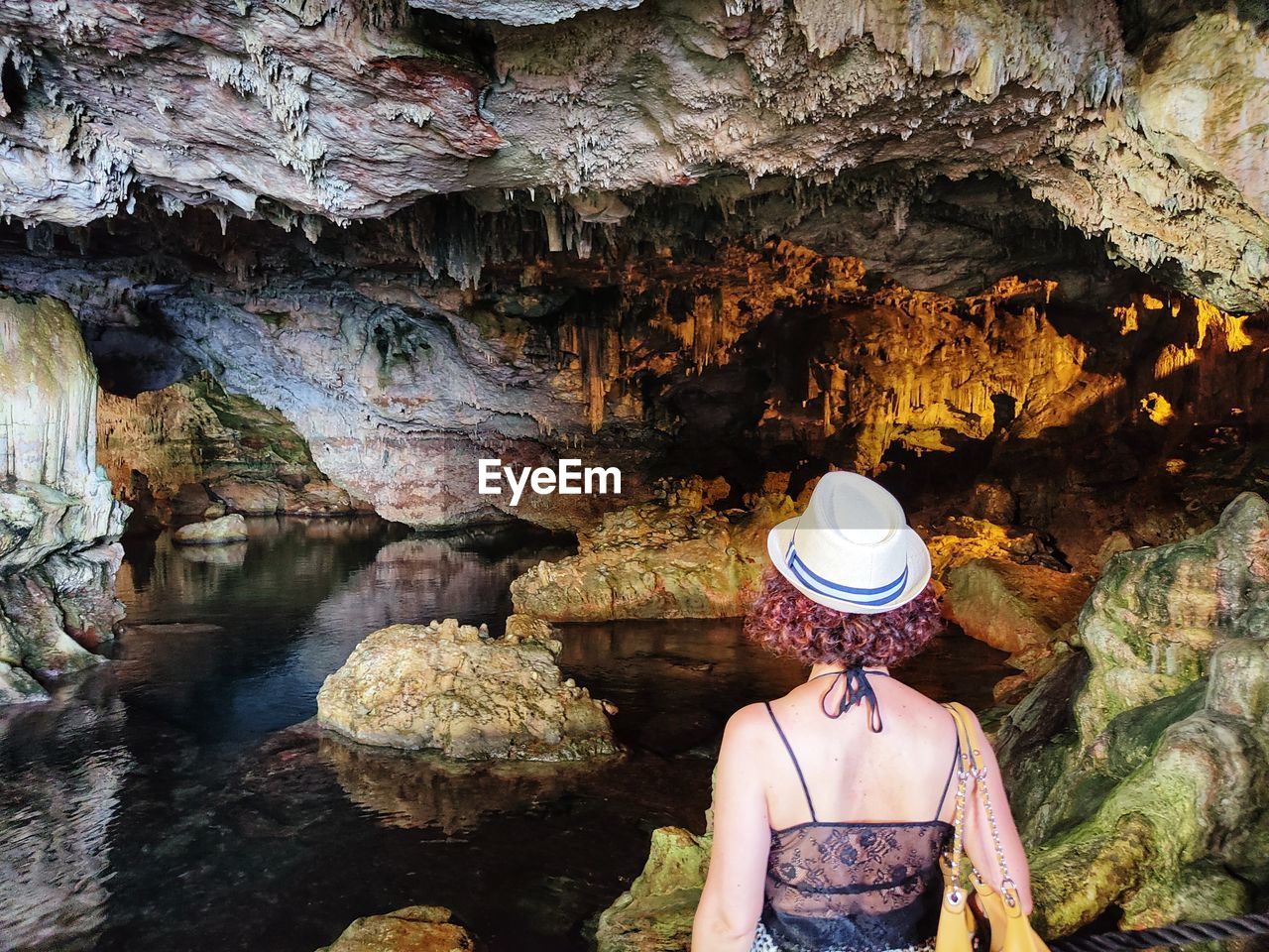 one person, rock, adult, hat, clothing, cave, nature, women, rock formation, caving, rear view, water, beauty in nature, travel, leisure activity, day, fashion accessory, formation, outdoors, lifestyles, geology, person, travel destinations, casual clothing, adventure, standing, tourism, sun hat, exploration, young adult