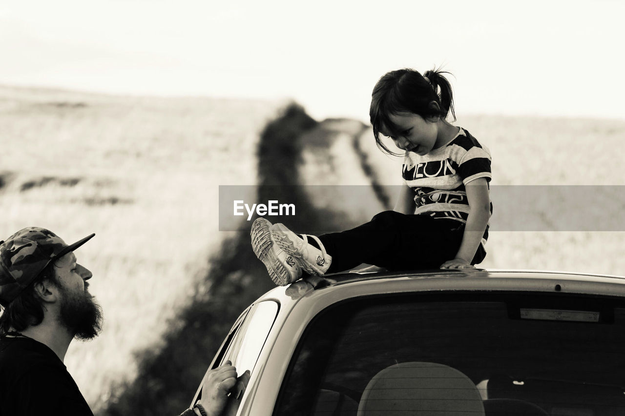 Side view of man looking at daughter sitting on car roof