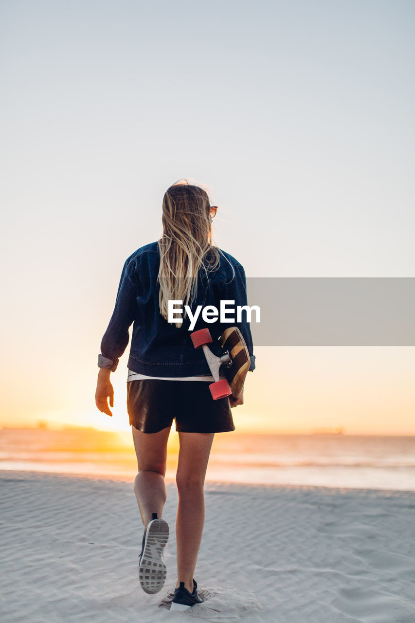 Rear view of young woman with skateboard walking at beach against clear sky during sunset