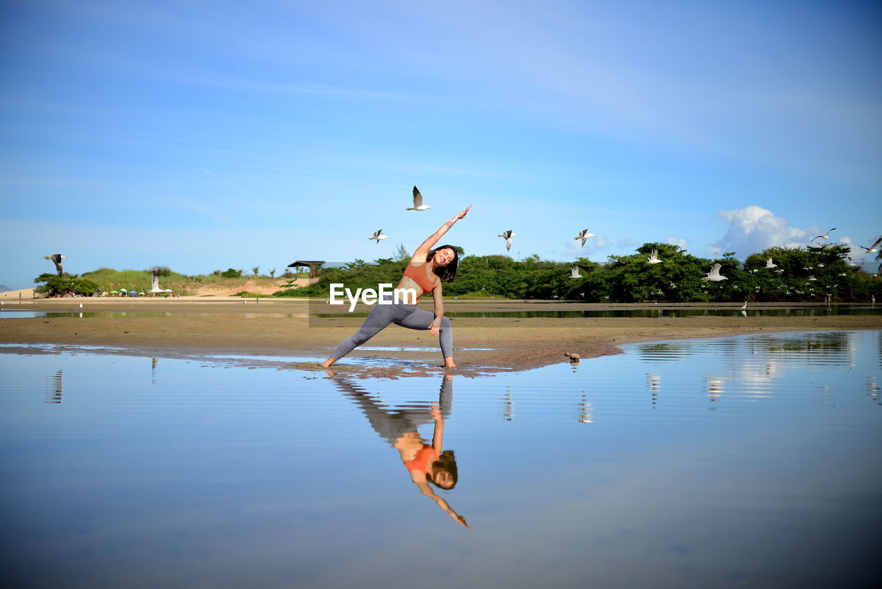 Young woman practicing yoga against blue sky, birds flying and her silhouette in the water.