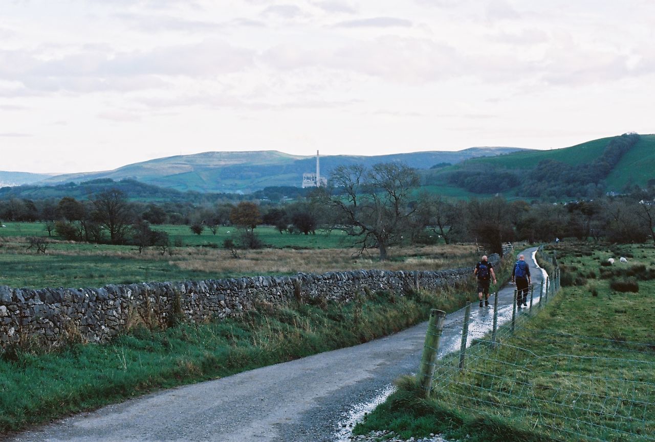 REAR VIEW OF PEOPLE WALKING ON ROAD AMIDST LANDSCAPE AGAINST SKY