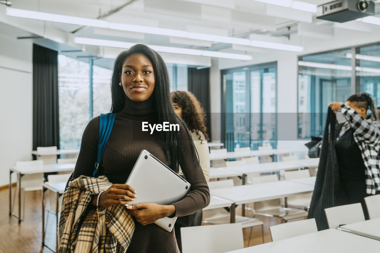 Portrait of smiling young woman holding jacket and laptop in classroom