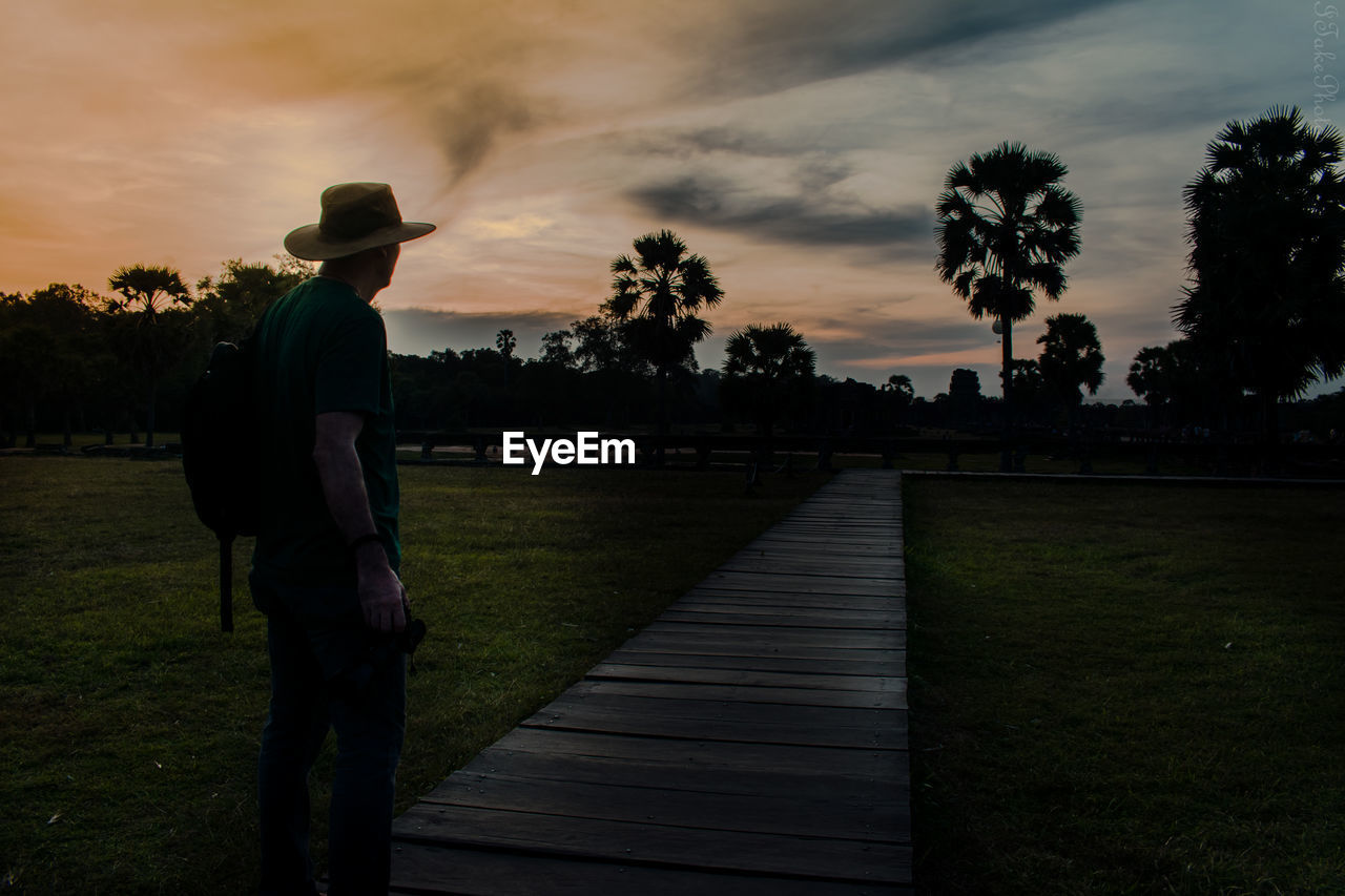 MAN STANDING BY FOOTPATH AGAINST SKY DURING SUNSET