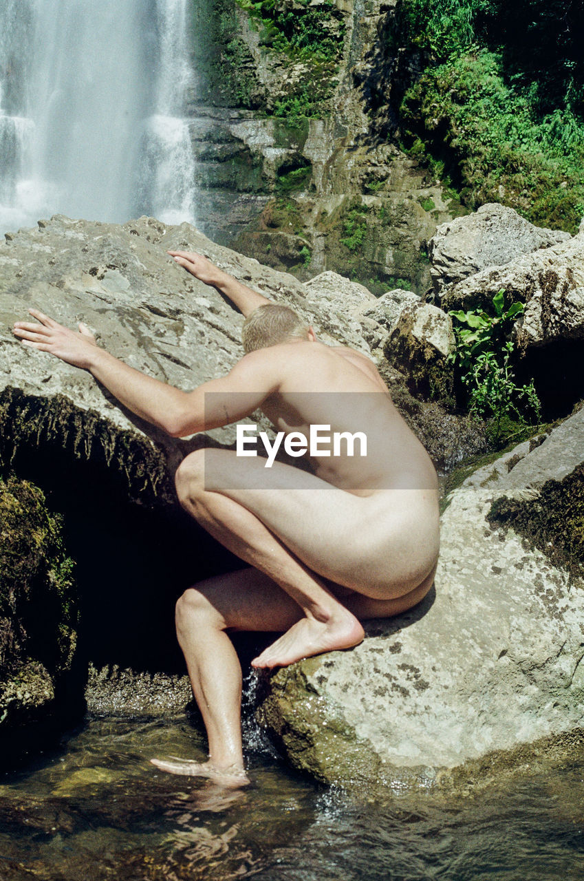 Full length of naked man sitting on rock against waterfall