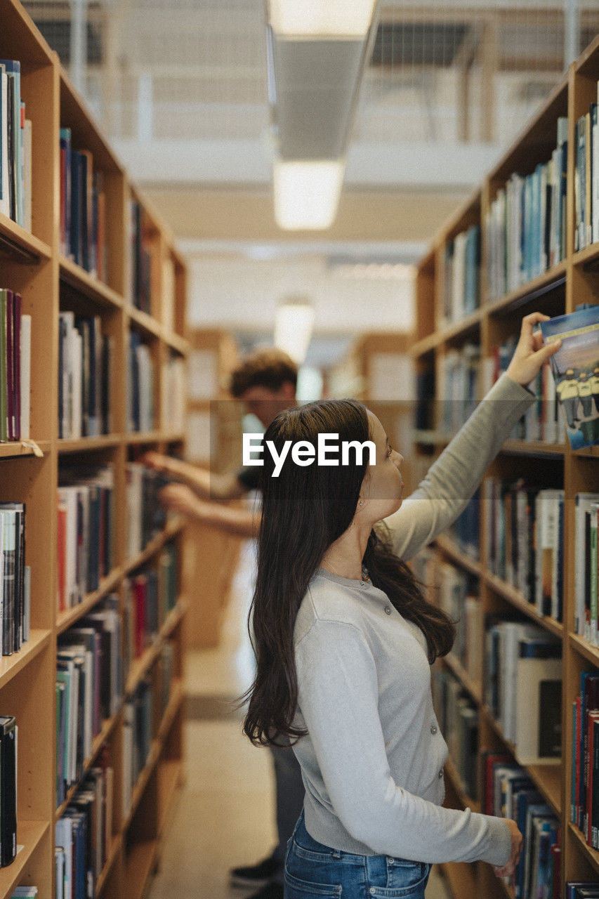 Side view of female student searching books on bookshelf in library at university