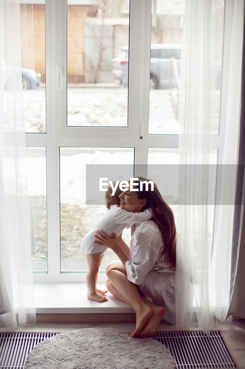 Mother in a white robe sits with a child a blonde daughter at a large window of the house person