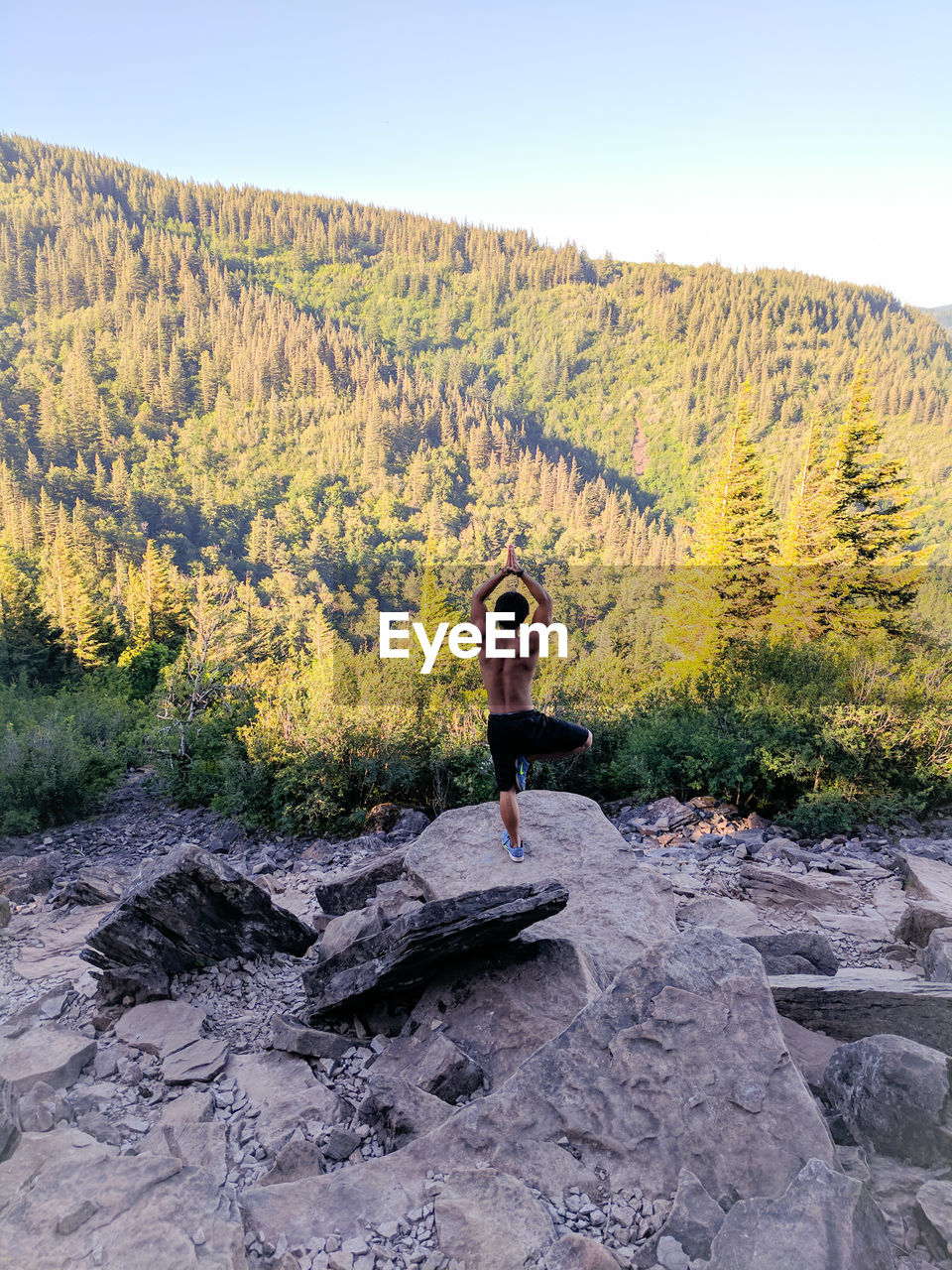 Rear view of young man exercising while standing on rock against sky