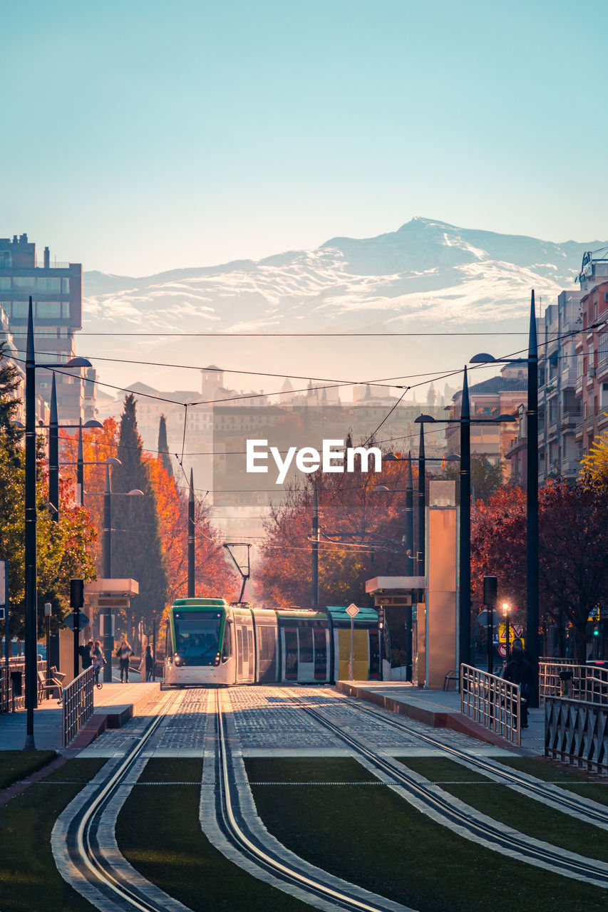 Contemporary tram driving on tracks near apartment buildings and deciduous trees against snowy mountain peak on autumn day in granada, spain