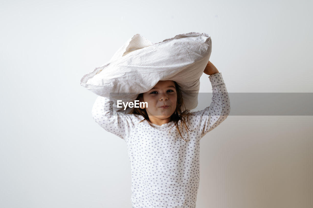 Portrait of cute girl holding pillow on head against wall