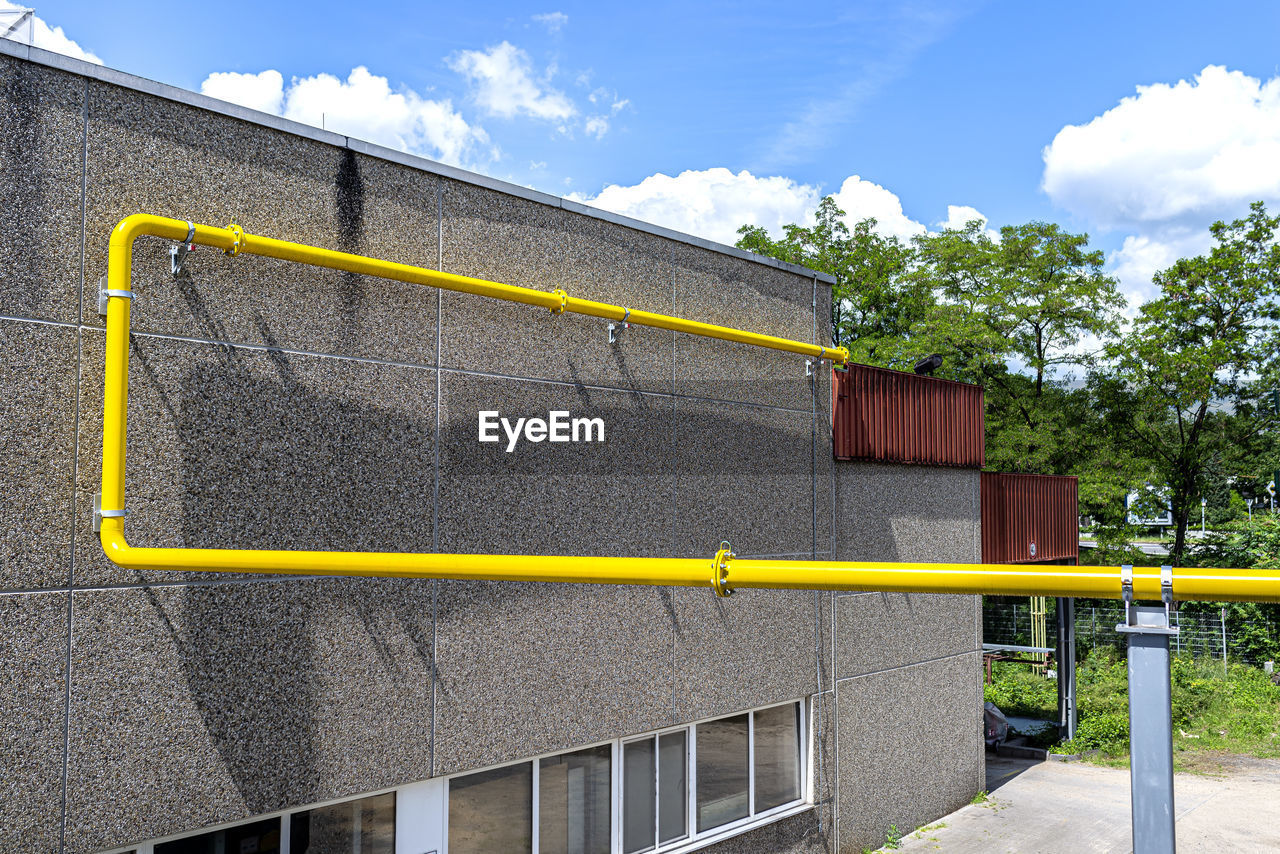 A large yellow gas pipe running up a factory wall made of concrete and pebbles.