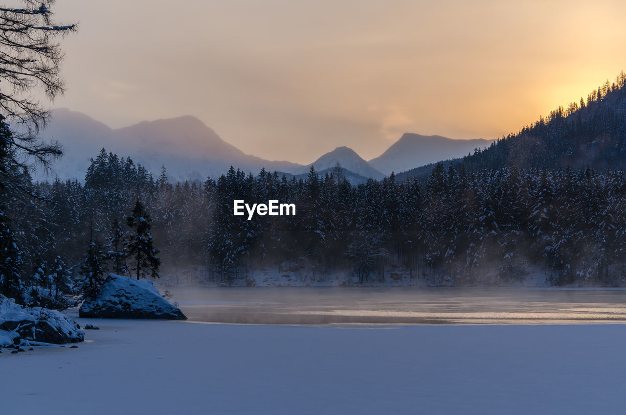 Frozen lake by trees against sky during sunrise