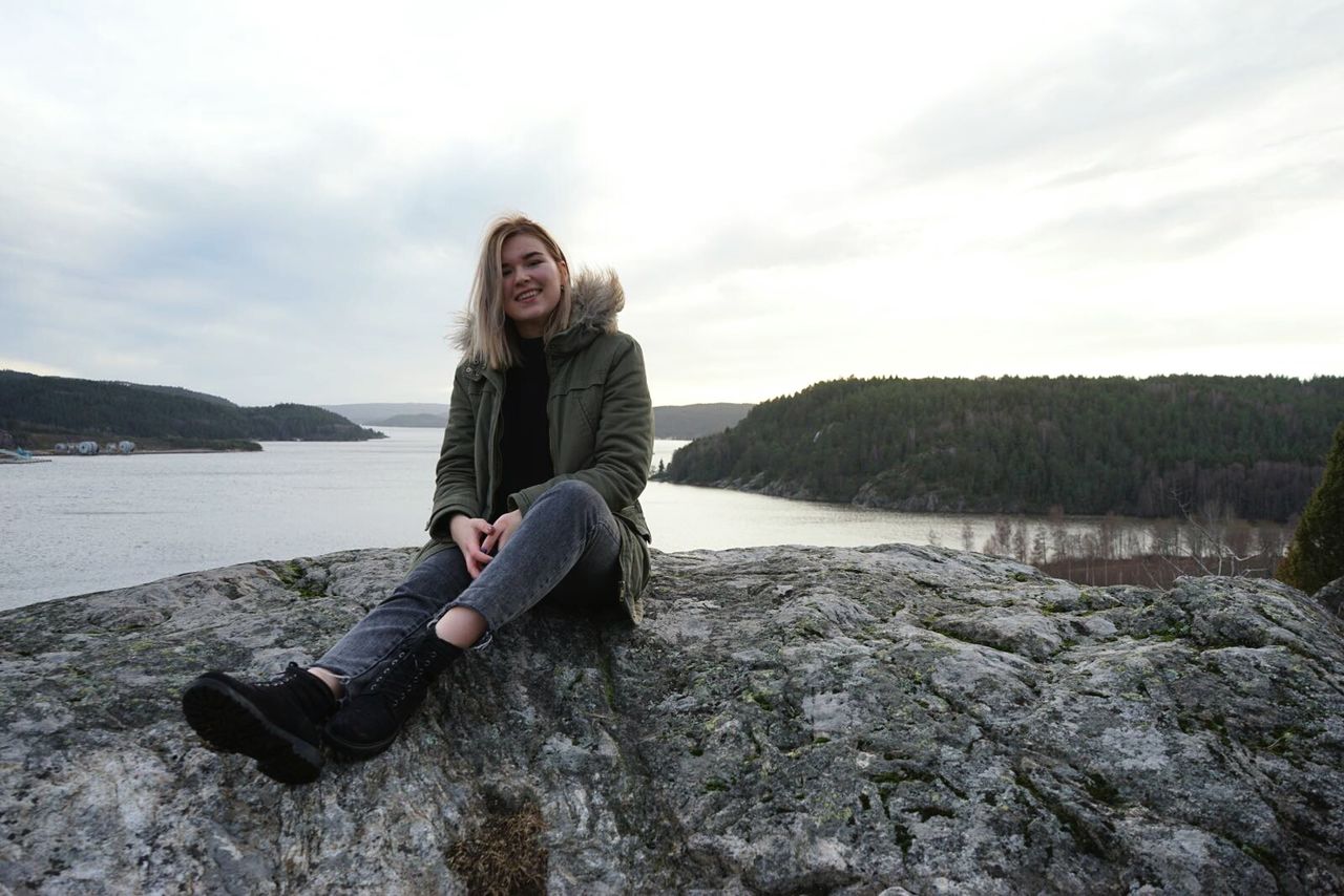 Full length of smiling woman sitting on rock by lake