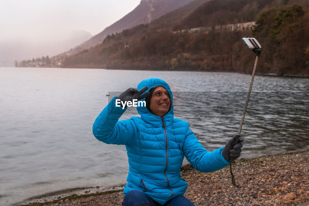 Woman in warm clothing taking selfie while sitting at lakeshore