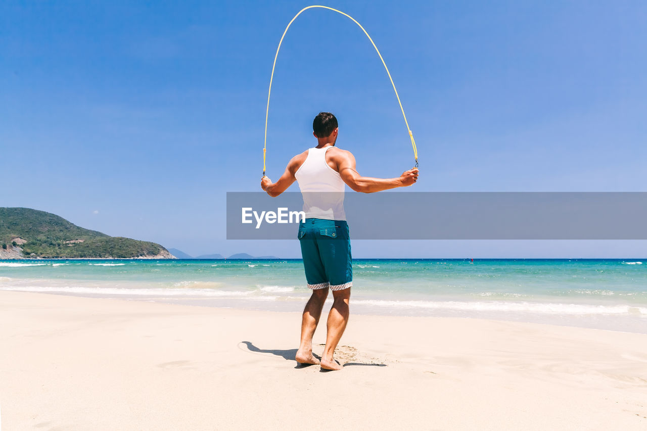 Rear view of man jumping rope at beach against blue sky