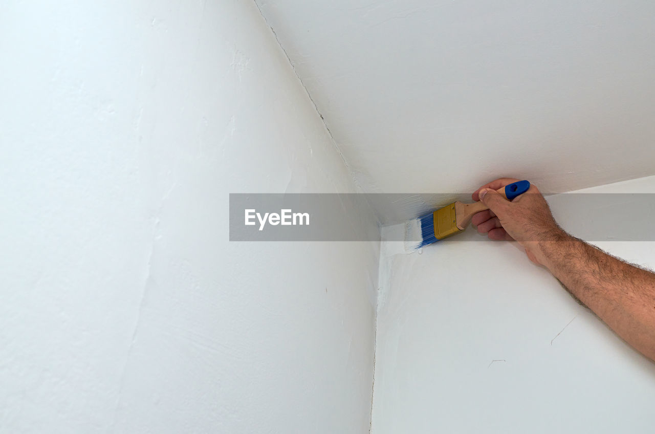 Hand holding paintbrush and painting corner of a wall and a ceiling