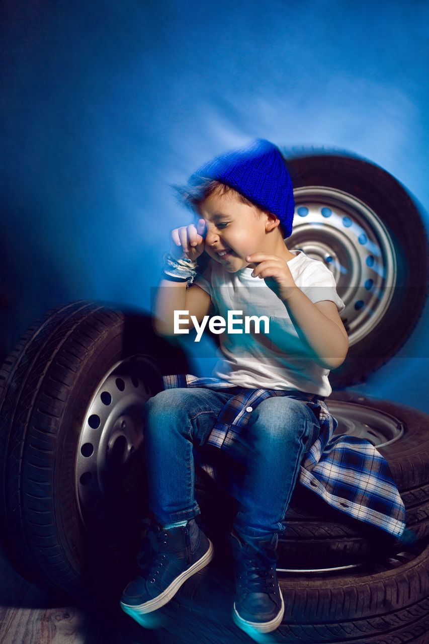 Boy in a white t shirt shirt and hat sits on a background of car wheels on a blue