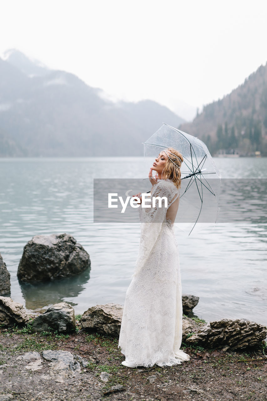 Beautiful young woman bride in a boho dress and with an umbrella stands in the rain in nature