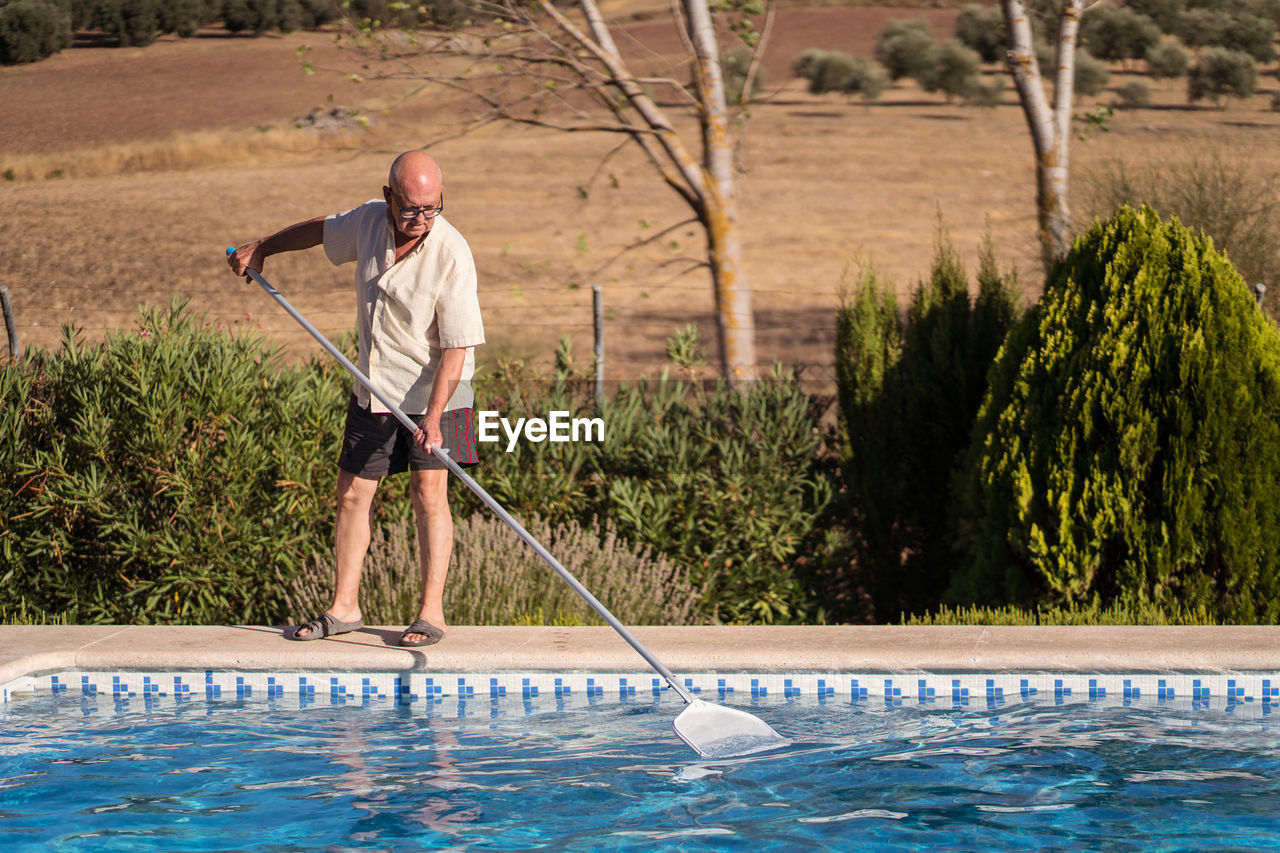 Full body of elderly male in eyeglasses cleaning water in swimming pool with mop in backyard