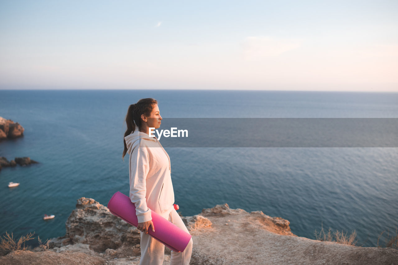 Smiling woman hold yoga mat wear sportswear hoodie and pants stand on top of rock over sea