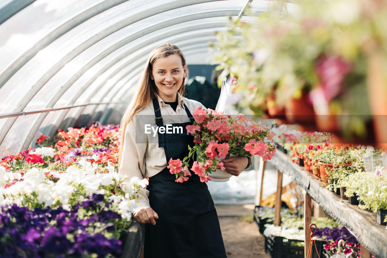 A cheerful woman in an apron holds a pot of flowers in her hands while working in a greenhouse. 