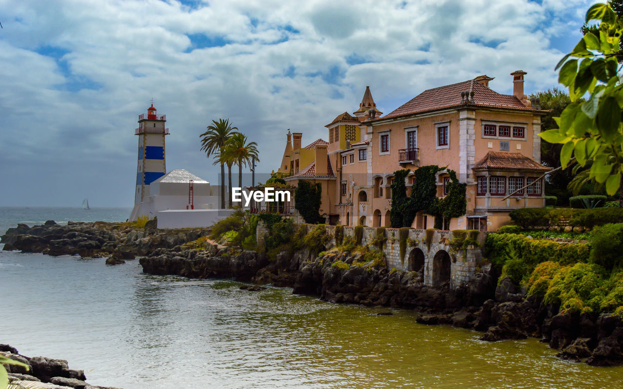 Lighthouse by sea and modern houses against sky