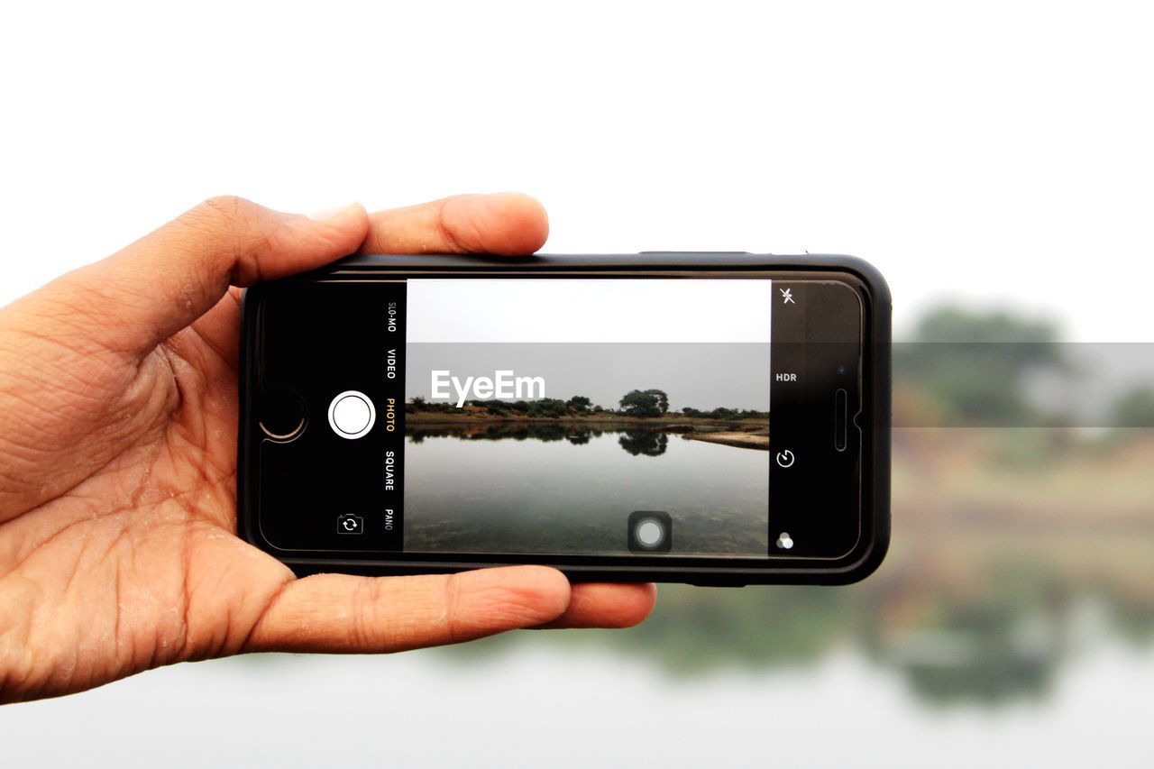 Cropped hand photographing lake against clear sky